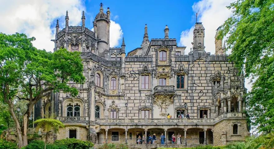 The Quinta da Regaleira estate in Sintra, Portugal, features a stunning palace with intricate stonework and ornate balconies, surrounded by lush greenery under a blue sky.