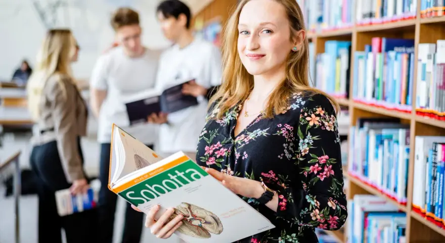 Studentka na Uniwersytecie Wrocławskim, kierunek prawo, przegląda atlas anatomii. W tle inni studenci i regały z książkami.