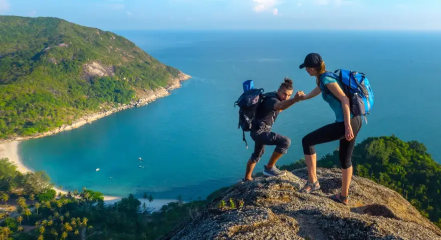 Due escursionisti si aiutano a salire su una scogliera con vista su una baia tropicale. Questo è il trekking cos'è: avventura e collaborazione.
