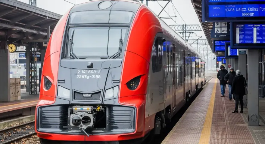 Czerwono-szary pociąg stoi na peronie, obok tablic informacyjnych z rozkładem jazdy.