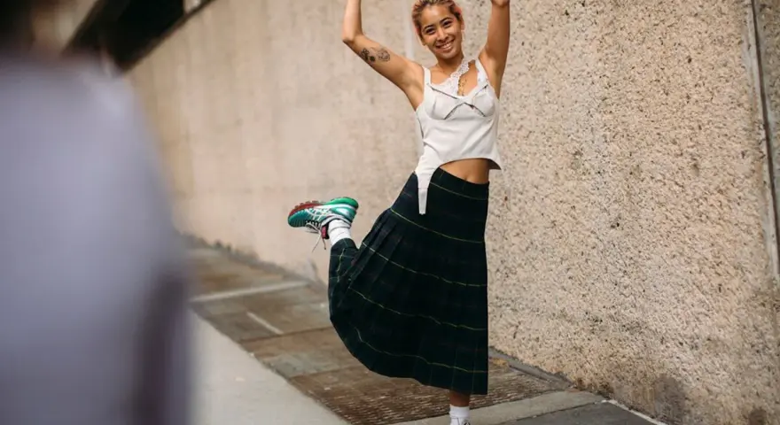 Joven sonriente con falda escocesa y top blanco, luciendo unos modernos looks con zapatillas deportivas.