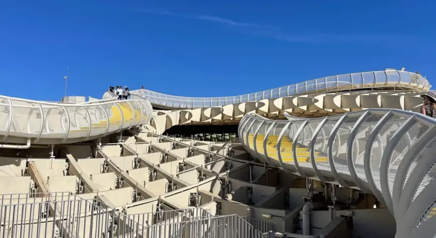 The Metropol Parasol, known as Las Setas de Sevilla, features a modern wooden structure with winding walkways and yellow steps under a clear blue sky.