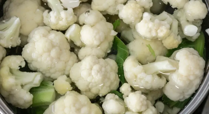 Kalafior gotuje się w garnku z wodą. Białe różyczki i zielone liście są zanurzone w płynie.