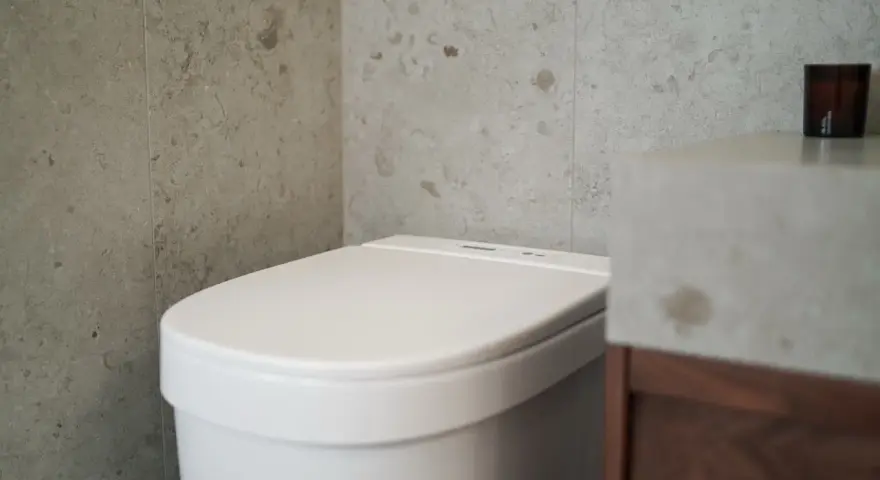 Modernt badrum med en vit toalett utan avlopp, grå kaklade väggar och ett handfat i betong.