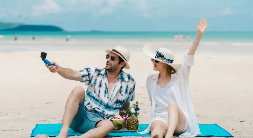 Para na plaży nagrywa filmik kamerką GoPro, a jego partnerka macha do obiektywu. Idealne wakacje pod gruszą!