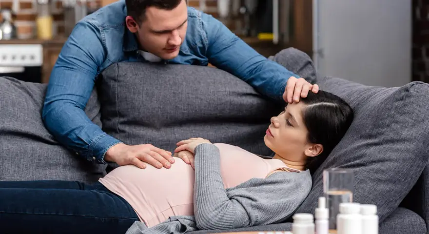 Zmartwiony partner troskliwie dotyka czoła ciężarnej kobiety, która leży na kanapie. Na stoliku leki, sugerujące problemy zdrowotne, być może angina w ciąży.