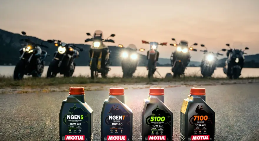 Cztery butelki oleju Motul do motocykla: NGEN5, NGEN7, 5100 i 7100, stoją na drodze o zachodzie słońca, za nimi widać rzędem motocykle.
