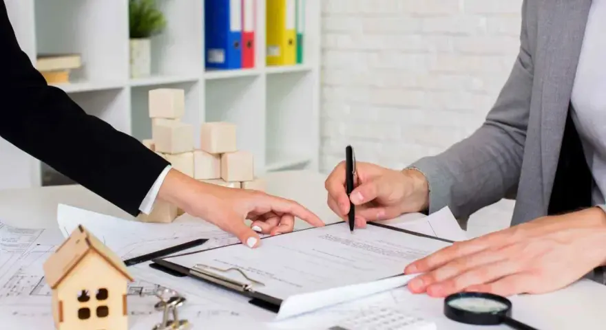 Podpisanie dokumentów, które mogą dotyczyć wypowiedzenia umowy zlecenia. Na stole leży model domu, klucze i lupa.