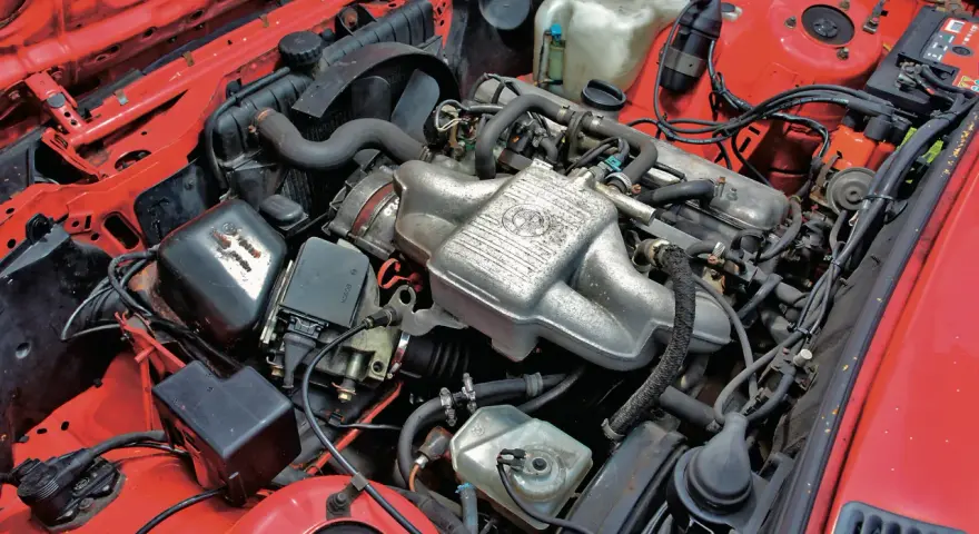 Silniki BMW E30 w czerwonej komorze silnika. Widoczny kolektor ssący z logo BMW, chłodnica, akumulator i inne elementy układu napędowego.