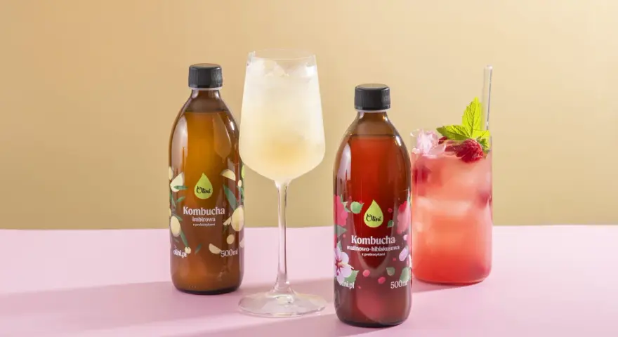 Kombucha co to? Dwie butelki kombuchy Olini: imbirowej i malinowo-hibiskusowej, obok orzeźwiających napojów z lodem.