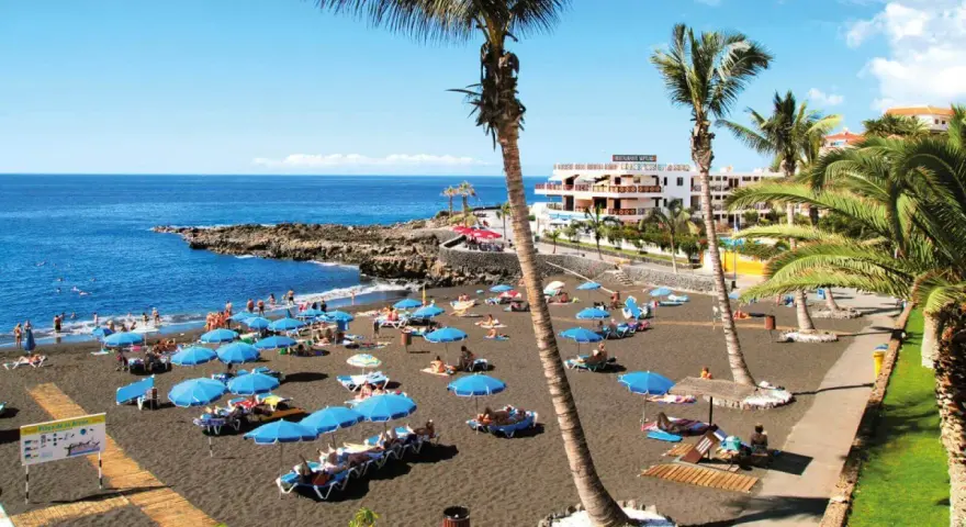 Czarna plaża Playa de la Arena na Teneryfie z turystami, niebieskimi parasolami i palmami.