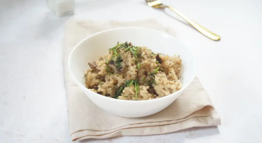 Miseczka kremowego risotto z grzybami, posypana ziołami i pieprzem, obok złoty widelec.