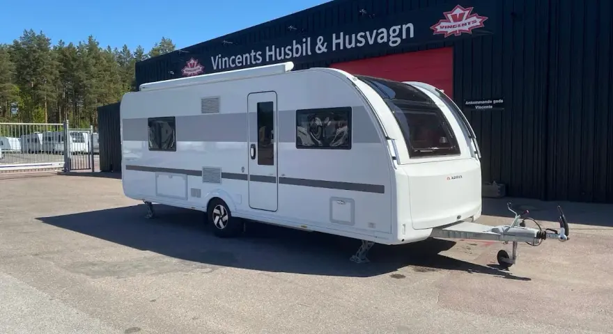 En vit Adria Alpina 613 UT husvagn står parkerad framför en byggnad med texten "Vincents Husbil & Husvagn".