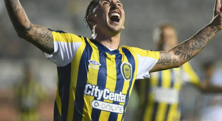 Jugador de Rosario Central celebra un gol, con los brazos en alto y la boca abierta. Sus tatuajes son visibles en sus brazos.