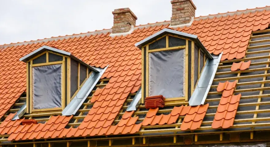 Trwa wymiana okna dachowego. Dach pokryty czerwona dachówką, widoczne drewniane elementy konstrukcji i nowe okna z folią.