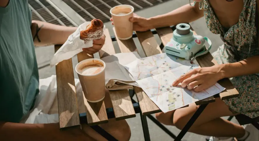 Para planuje podróż, studiując mapę i jedząc croissanty. Na stole kawa i aparat.