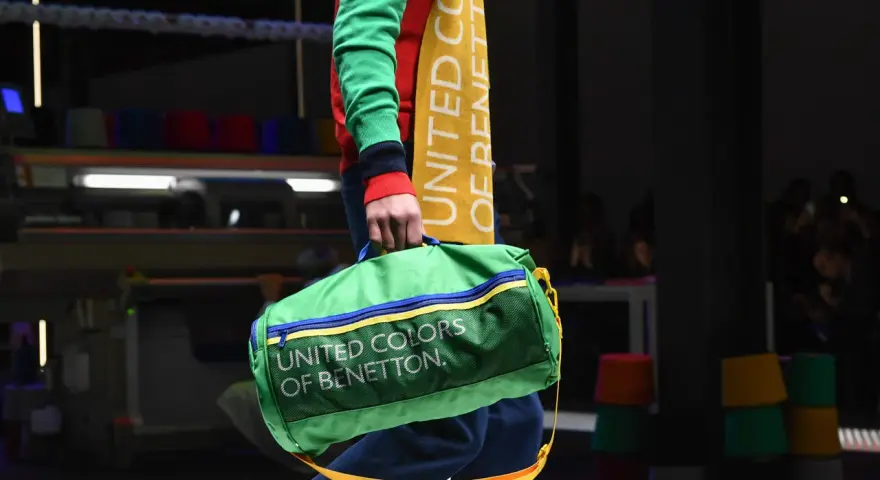 Zielony worek z napisem "UNITED COLORS OF BENETTON" niesiony przez modela w kolorowym swetrze.