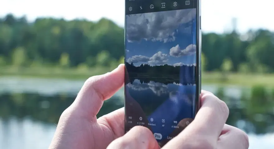 Dłonie trzymają smartfon Samsung S10, na ekranie którego widać odbicie nieba i lasu w spokojnej wodzie.