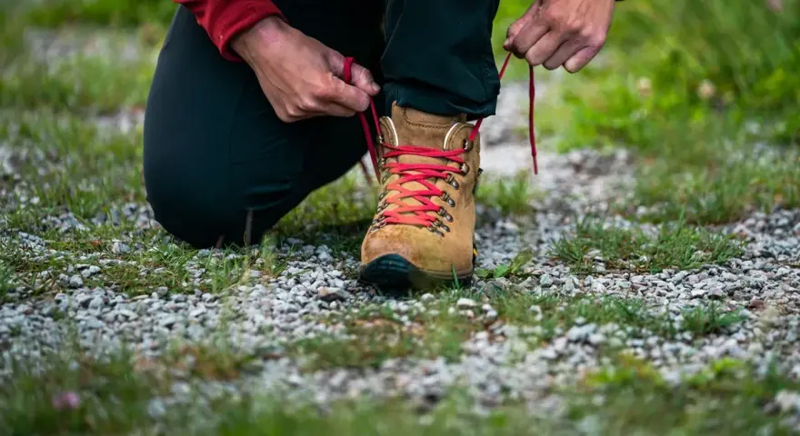 Osoba klęczy i sznuruje brązowy but trekkingowy z czerwonymi sznurówkami. To dobry przykład, jak zmierzyć stopę przed zakupem obuwia.