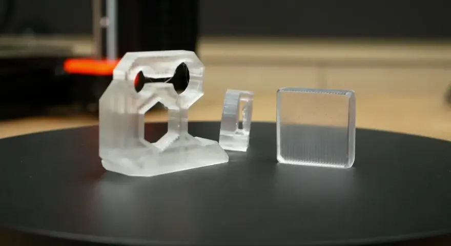Three clear 3D printed objects, one complex bracket and two simpler shapes, sit on a black surface.