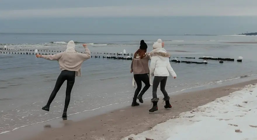 Trzy dziewczyny cieszą się zimowym dniem, gdzie zimą nad morze. Spacerują po plaży, na której leży śnieg, a fale delikatnie rozbijają się o brzeg.