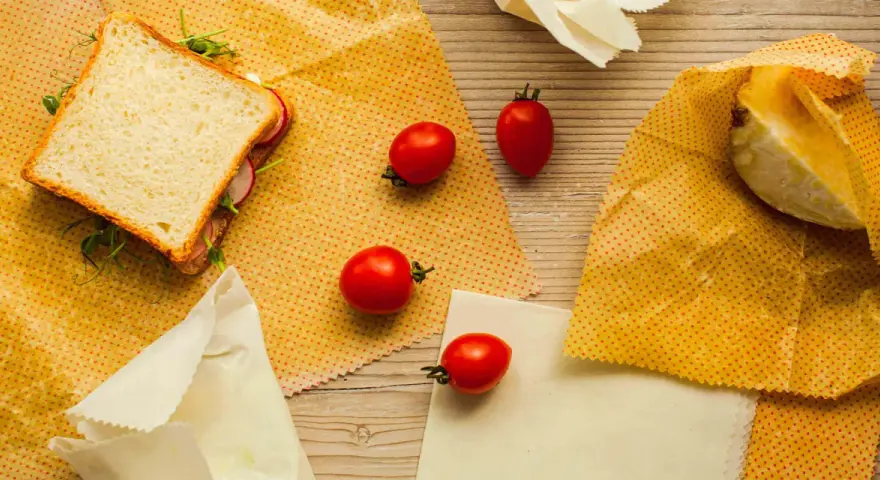 Un sandwich, des tomates et un morceau de fromage sont présentés sur des bee wraps maison à pois rouges sur fond de bois.