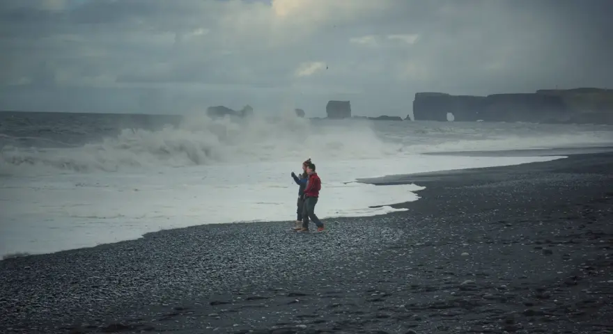 Para spaceruje po czarnej plaży Islandii, podczas gdy fale rozbijają się o brzeg.