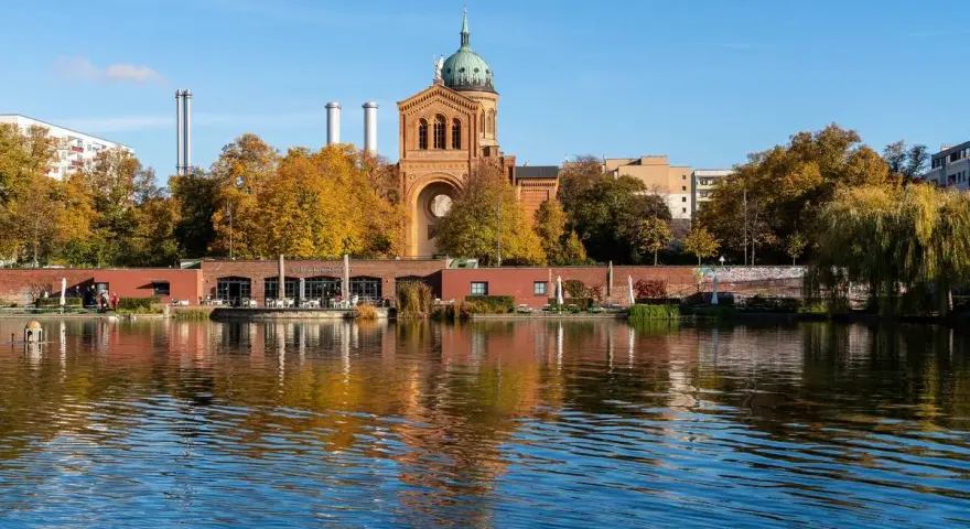 Herbstliche Bäume spiegeln sich im Wasser des Engelbeckens Berlin. Ein Café mit Außenbereich und eine Kirche mit Kuppel sind zu sehen.