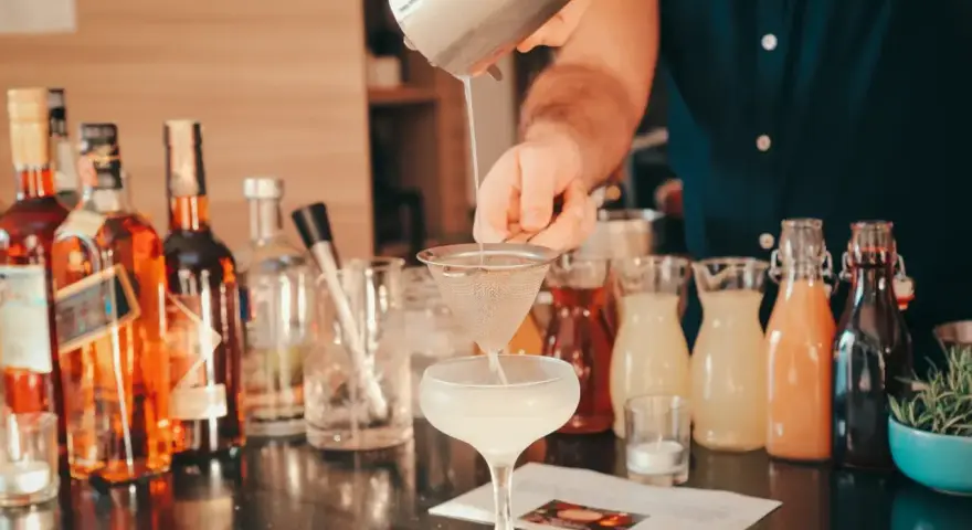 Barman przelewa płyn z shakera przez sitko do kieliszka, tworząc koktajl. W tle butelki alkoholi i kolorowe syropy.