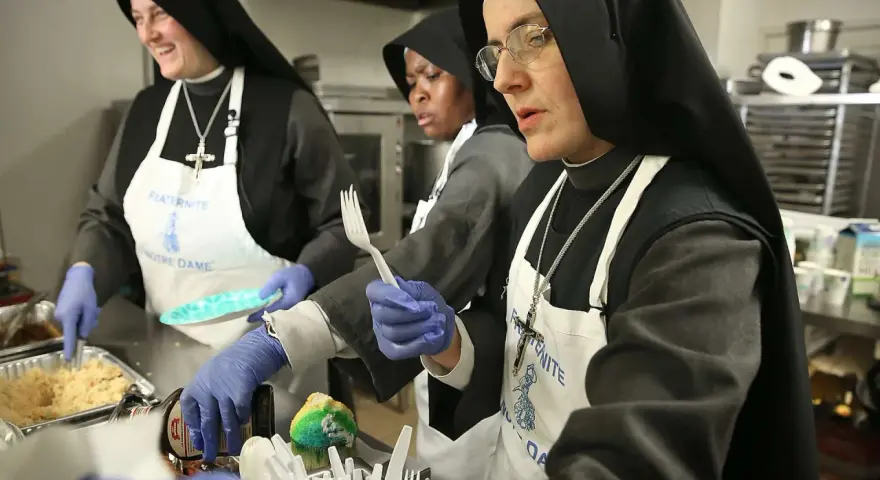 Nuns in a kitchen preparing food. One nun smiles while serving, another holds a fork, and a third looks on. This scene might answer the question: do nuns eat meat?