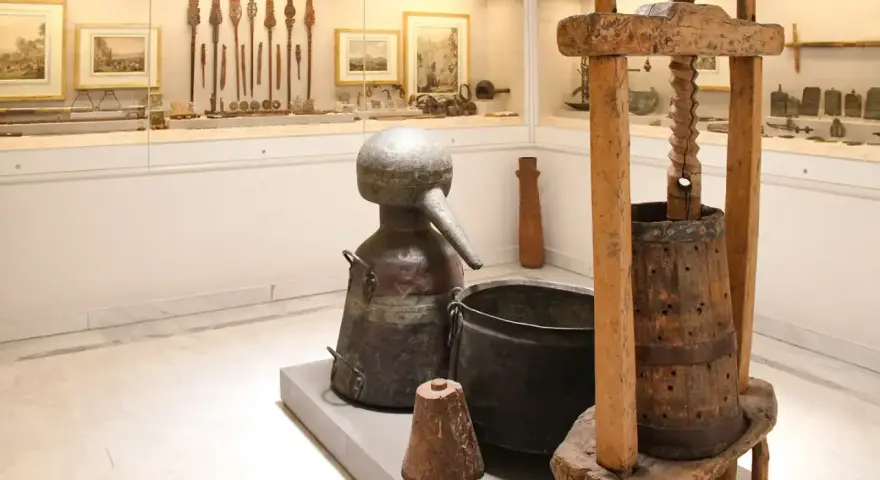 W muzeum Ateny: zabytkowy alembik, kadź i prasa do winogron. W tle ekspozycja narzędzi i obrazów.