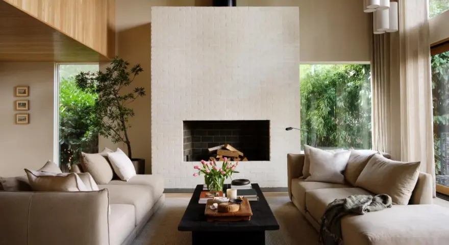Salón de estilo japandi con sofás beige, chimenea de ladrillo blanco y mucha luz natural.