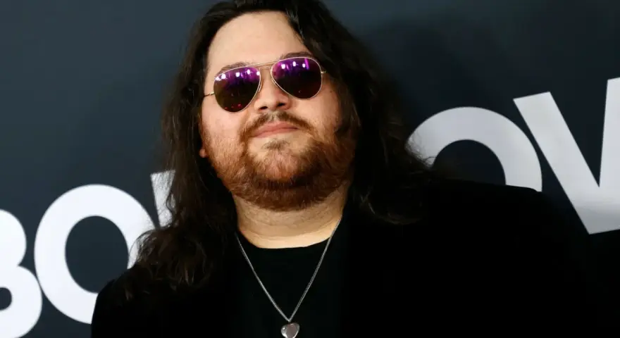 Wolfgang Van Halen w okularach przeciwsłonecznych i z naszyjnikiem w kształcie serca, na tle logo HBO.