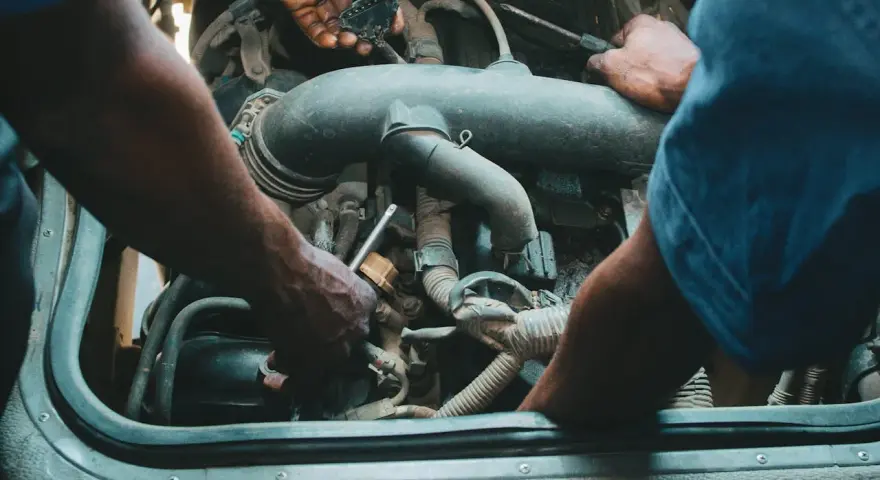Mechanicy pracują przy silniku, jeden z nich trzyma wtyczkę, drugi dokręca śrubę. Może to być naprawa zaworu par paliwa.