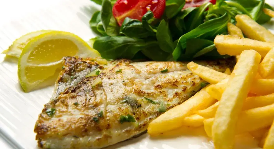 A cooked fish fillet with french fries, salad, and lemon slices on a white plate.