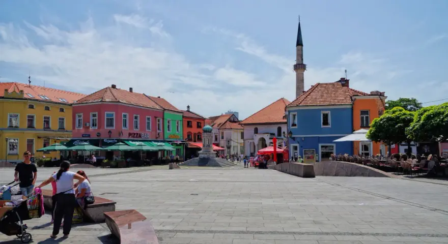 Kolorowy plac w **Tuzla, Bośnia**, z kawiarniami, fontanną i minaretem w tle.