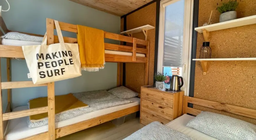 Wnętrze chałupy kempingowej z łóżkiem piętrowym i pojedynczym, żółtym kocem i torbą z napisem "MAKING PEOPLE SURF".