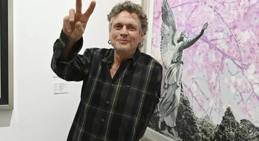Rick Allen, perkusista Def Leppard, pozuje z uniesionym znakiem pokoju obok obrazu z aniołem i kwitnącymi drzewami.