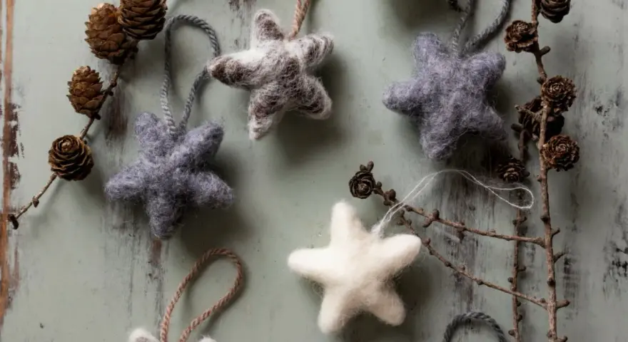 Mjuka, filtade julstjärnor i grått och vitt, som påminner om klassiska julpyssel. Dekorerade med kottar och grenar på en sliten träbakgrund.