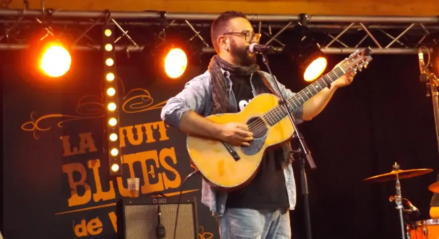 Un homme joue de la guitare sur scène, éclairé par des projecteurs. Le panneau derrière lui annonce "La Nuit du Blues", évoquant la naissance du blues.