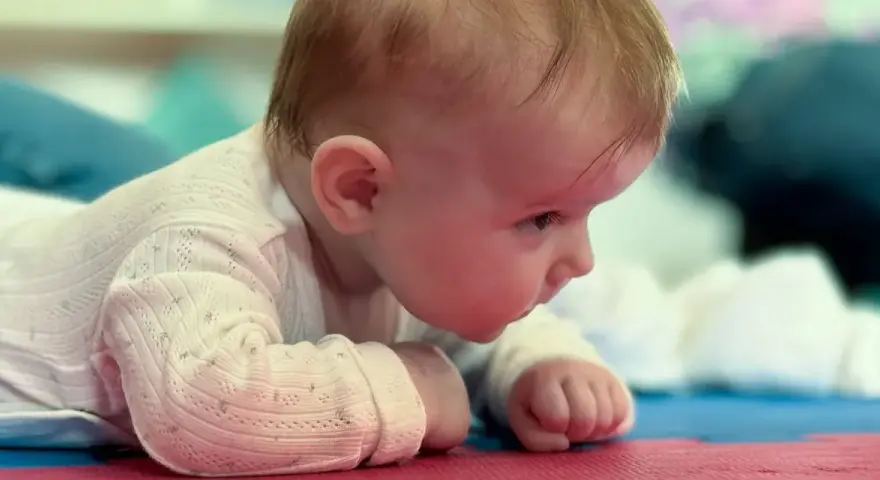 Malutkie dziecko leży na brzuchu, z zaciekawieniem spoglądając przed siebie. Jego duża głowa u dziecka jest lekko pochylona, a drobne rączki podparte o podłoże.