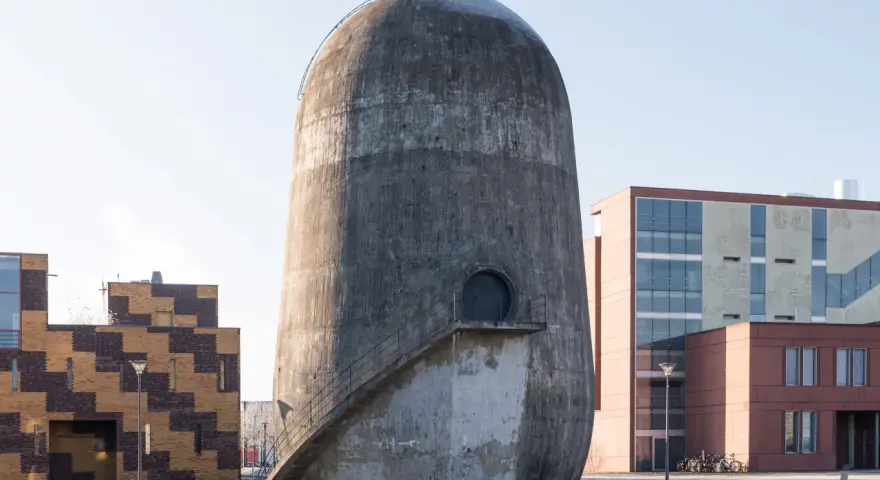 Der Trudelturm Berlin, ein futuristisches Betonbauwerk, steht neben modernen Gebäuden. Eine Außentreppe führt zu einer runden Tür.