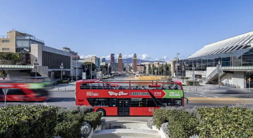 A red double-decker bus, the best hop on hop off Barcelona, drives past modern architecture under a clear blue sky.