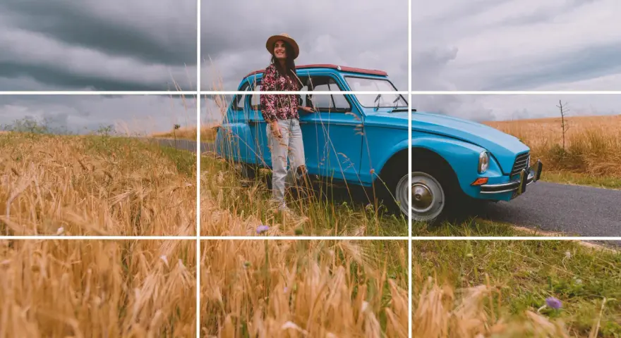 Une femme souriante, chapeau de paille, pose près d'une voiture bleue vintage au milieu d'un champ de blé doré. Photos prises sur le vif.