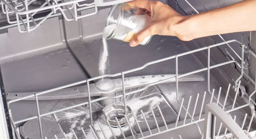 A hand pours white powder from a jar into an open dishwasher, demonstrating how to clean a dishwasher naturally.