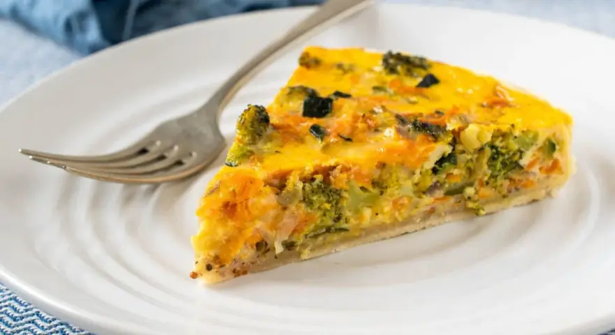 Rebanada de quiche vegetal con brócoli y zanahoria, servida en plato blanco con tenedor.