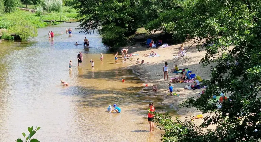 Słoneczny dzień nad Świdrem. Ludzie odpoczywają na piaszczystej plaży i kąpią się w rzece. Dzieci bawią się w wodzie.
