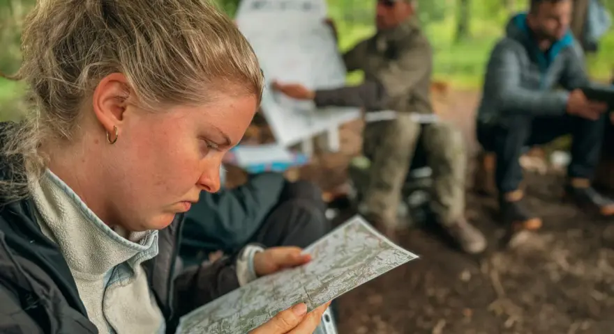 Frau studiert Karte, während Männer im Hintergrund bei einer Ausbildung im Wald sitzen. Solche intensiven, kurzen **ausbildungen die nur 1 jahr dauern** sind ideal für schnelle Karriereschritte.