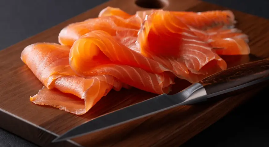 Finas lonchas de salmón ahumado, listas para disfrutar, reposan sobre una tabla de madera junto a un cuchillo.