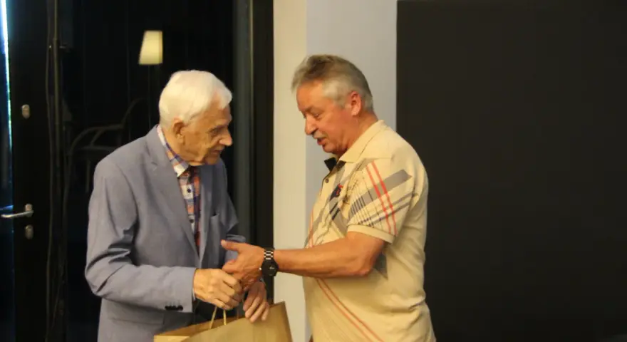Zdzisław Kuźniar syn, starszy pan w niebieskiej marynarce, wręcza prezent młodszemu mężczyźnie w beżowej koszulce.
